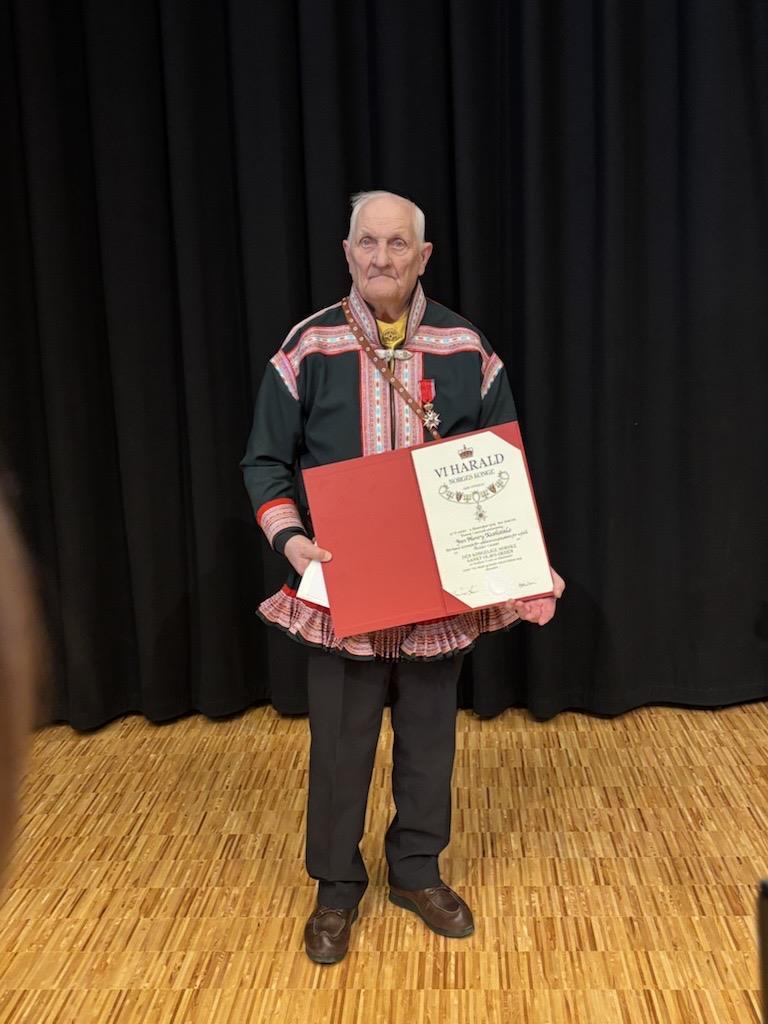 Jan Henry Keskitalo viser fram diplomet som viser at han er mottaker Den Kongelige Norske St. Olavs Orden. Han har mørk kautokeinokofte på seg, i bakgrunnen svart sceneteppe - Deaddil ja stuora govva ihtá