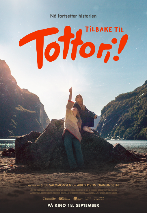 Kinoplakat for filmen «Tilbake til Tottori!». To jenter står ved en stor stein på en strand omgitt av høye fjell og en fjord. Den ene jenta peker opp mot himmelen. Øverst på plakaten står teksten «Nå fortsetter historien» og filmens tittel i store, oransjerøde bokstaver. Nederst står det «På kino 18. september»